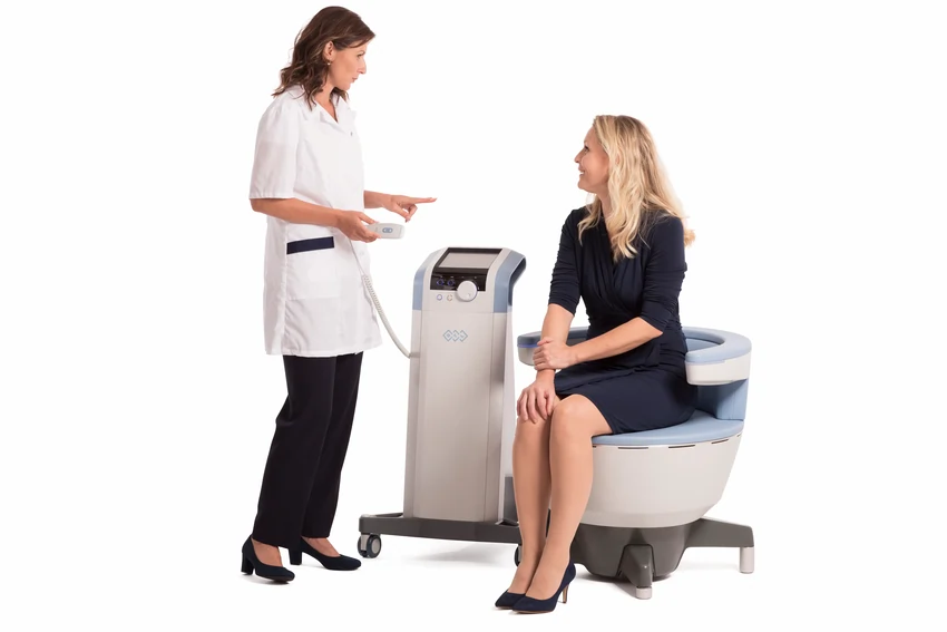 Pelvic floor specialist explaining Emsella treatment to a seated client, with the Emsella machine in the middle, demonstrating non-invasive therapy